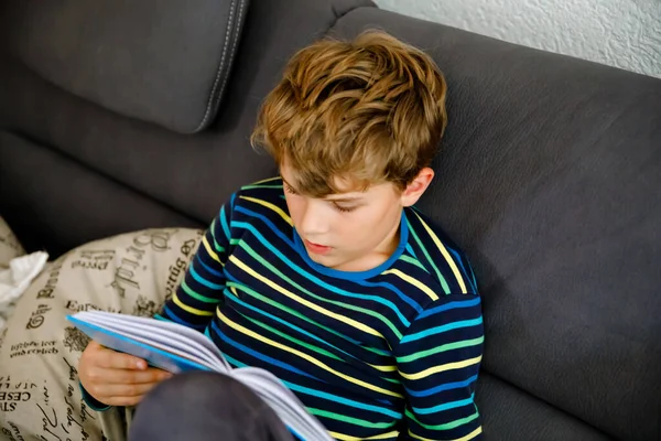 Lindo niño rubio pequeño leyendo revista o libro en la habitación doméstica. Un niño emocionado leyendo alto, sentado en el sofá. Colegial, familia, educación. Educación en el hogar durante el tiempo covid — Foto de Stock