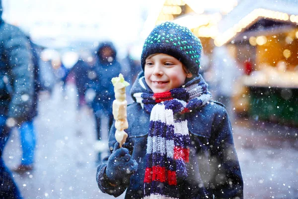 Petit garçon mignon mangeant des fruits couverts de chocolat blanc sur brochette sur le marché de Noël allemand traditionnel. Enfant heureux sur le marché familial traditionnel en Allemagne pendant la journée enneigée. — Photo