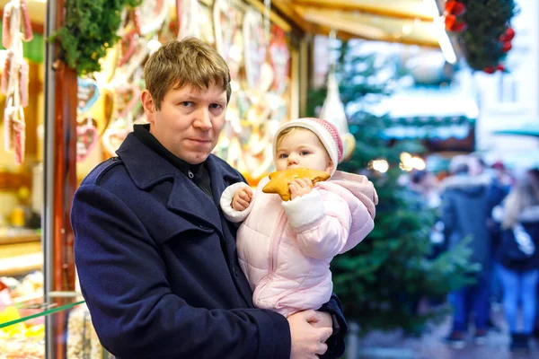 Отец средних лет держит дочку рядом со сладким ларьком с пряниками и орехами. Счастливая семья на рождественском рынке в Германии. Милая девушка ест печенье по имени Лебкучен. Празднование Рождества Христова. — стоковое фото