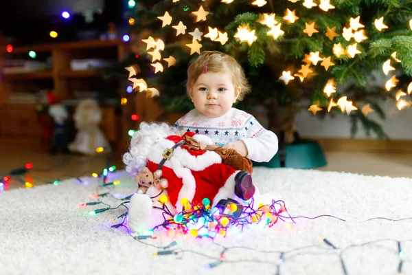 可愛い手でカラフルなライトガーランドを保持する愛らしい赤ちゃんの女の子。クリスマスツリーを家族で飾るお祝い服の小さな子供。Weihnachtenと呼ばれる伝統的な休日の最初のお祝い — ストック写真