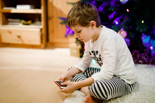 Söt liten blond pojke med hjälp av smartphone på jul med dekorerade träd på bakgrunden. Glad hälsosam hild ha kul hemma. Populär present till jul — Stockfoto