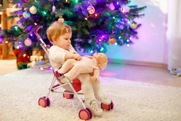 Cute adorable baby girl having fun with with with doll carriage. Ребенок сидит дома в коляске с игрушкой. Счастливая дочь. С ёлкой и огнями. — стоковое фото