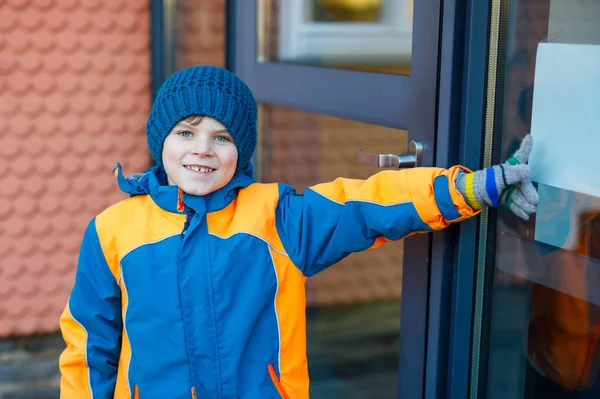 寒い冬の日に小学校の子供の男の子が学校に歩いています。学校への扉を押す暖かい冬の服の幸せな子供、屋外. — ストック写真