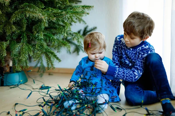 Cute baby girl and kid boy taking down holiday decorations from Christmas tree. children, siblings, brother and sister holding light garland. Family after celebration remove and dispose tree — Stock Photo, Image