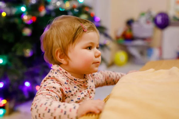 Πορτρέτο του μικρού χαριτωμένο κοριτσάκι μάθηση περπάτημα και όρθια. με χριστουγεννιάτικο δέντρο και φώτα στο παρασκήνιο. Αξιολάτρευτο κοριτσάκι στο σπίτι. Όμορφο μωρό με καθημερινά ρούχα. — Φωτογραφία Αρχείου