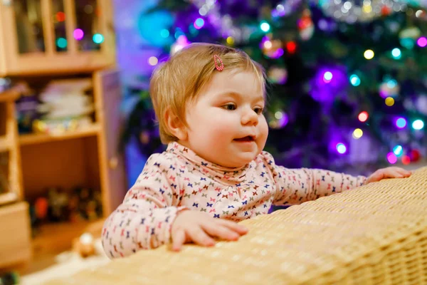 Portrait of little cute baby girl learning walking and standing. with Chrismas tree and lights on background. Adorable toddler girl at home. Beautiful baby child in casual clothes — Stock Photo, Image
