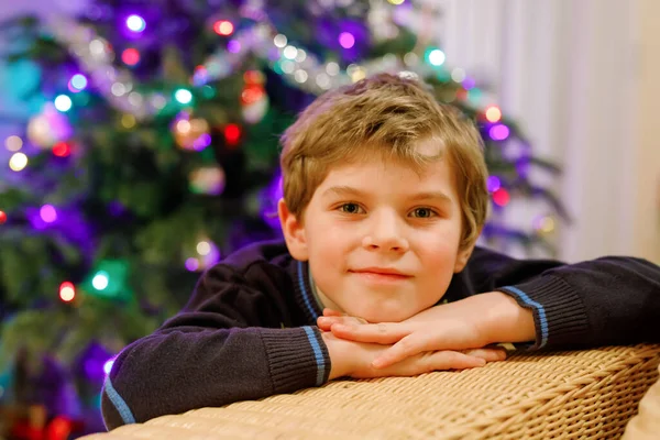 Kleiner Junge an Heiligabend mit Weihnachtsbaum und Lichtern im Hintergrund. Mit farbenfroher Beleuchtung und Girlanden. Glückliches Vorschulkind, das Weihnachten feiert, Familienurlaub. Junge in Nachthemd. — Stockfoto