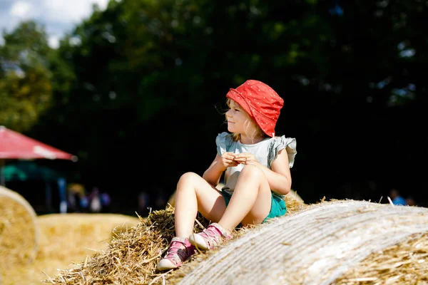 Pequeña niña divirtiéndose con correr y saltar en la pila de heno o paca. Divertido niño feliz y saludable jugando con paja. Activo ocio al aire libre con los niños en el cálido día de verano. Niños y naturaleza —  Fotos de Stock