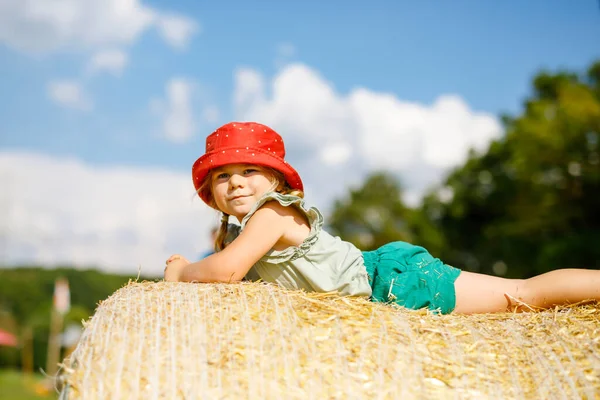 Маленькая девочка развлекается бегом и прыжками на стоге сена или тюке. Смешной счастливый здоровый ребенок играет с соломой. Активный отдых на свежем воздухе с детьми в теплый летний день. Дети и природа — стоковое фото