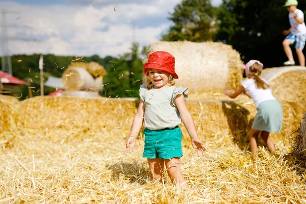 小さな幼児の女の子は、干し草の山やベールで走ったりジャンプしたりして楽しんでいます。藁で遊ぶ面白い幸せな健康な子供。暖かい夏の日に子供たちと積極的な屋外レジャー。子供と自然 — ストック写真