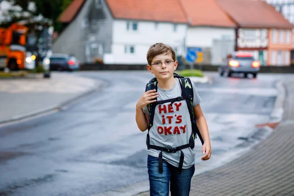 Happy little kid boy with glasses and backpack or satchel. Schoolkid on the way to middle or high school. Healthy adorable child outdoors on the street, on rainy day. Back to school. — Stock Photo, Image