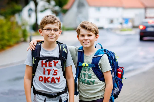 Dos niños pequeños con mochila o mochila. Los colegiales de camino a la escuela. Niños sanos y adorables, hermanos y mejores amigos al aire libre en la calle saliendo de casa. De vuelta a la escuela. Felices hermanos.. — Foto de Stock