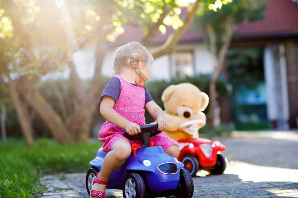 Pequeña niña adorable conduciendo un coche de juguete y divirtiéndose jugando con un oso de peluche, al aire libre. Precioso niño feliz y saludable disfrutando de un cálido día de verano. Sonriente niño impresionante en gaden —  Fotos de Stock