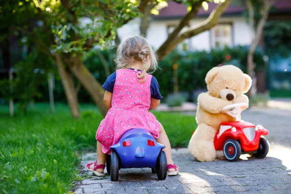 Pequeña niña adorable conduciendo un coche de juguete y divirtiéndose jugando con un oso de peluche, al aire libre. Precioso niño feliz y saludable disfrutando de un cálido día de verano. Sonriente niño impresionante en gaden —  Fotos de Stock