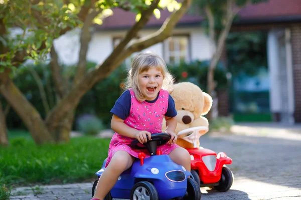 Malý rozkošný batole dívka řízení autíčko a baví se hrát s plyšovým medvídkem, venku. Nádherné, šťastné a zdravé dítě si užívá teplého letního dne. Usmívající se ohromující dítě v gadenu — Stock fotografie