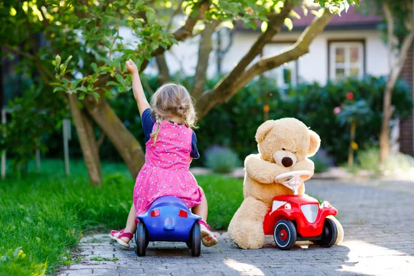 Pequena menina adorável criança dirigindo carro de brinquedo e se divertindo com brincar com urso de brinquedo de pelúcia, ao ar livre. Linda criança saudável feliz desfrutando de dia quente de verão. Sorrindo garoto deslumbrante em gaden — Fotografia de Stock