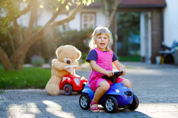 Pequeña niña adorable conduciendo un coche de juguete y divirtiéndose jugando con un oso de peluche, al aire libre. Precioso niño feliz y saludable disfrutando de un cálido día de verano. Sonriente niño impresionante en gaden — Foto de Stock