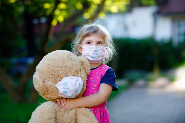 Little toddler girl in medical mask as protection against pandemic coronavirus quarantine disease. Cute child using protective equipment as fight against covid 19 and holding big bear toy with mask.