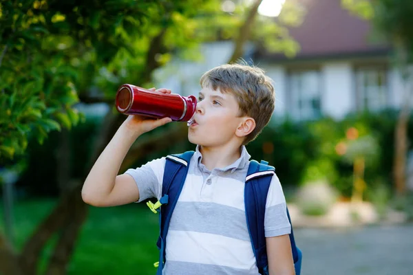 Счастливый маленький мальчик пьет из бутылки с водой, рюкзака или сумки. Школьник на пути в школу. Здоровый ребенок на улице. Возвращение в школу. — стоковое фото
