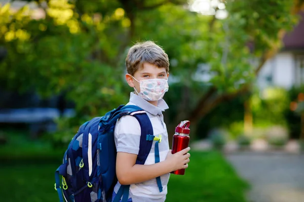 Rapazinho feliz, máscara médica, garrafa de água e mochila ou mochila. Um estudante a caminho da escola. Criança saudável ao ar livre. De volta à escola após o tempo de quarentena do bloqueio da doença pandêmica da corona — Fotografia de Stock