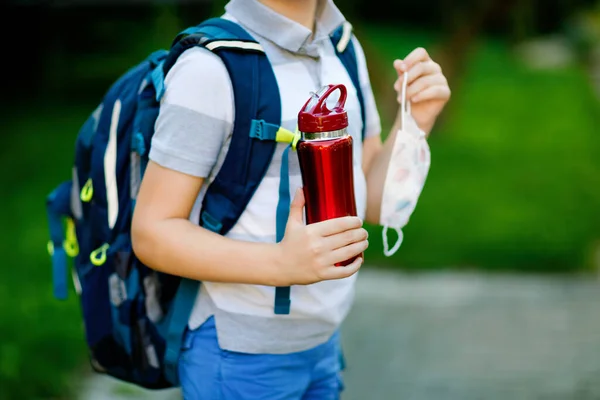 子供の男の子、医療用マスク、水ボトルやバックパックやサッチェルの閉鎖。学校に行く途中の小学生。屋外の子供。コロナパンデミックの予防接種後、学校に戻る。顔もない. — ストック写真