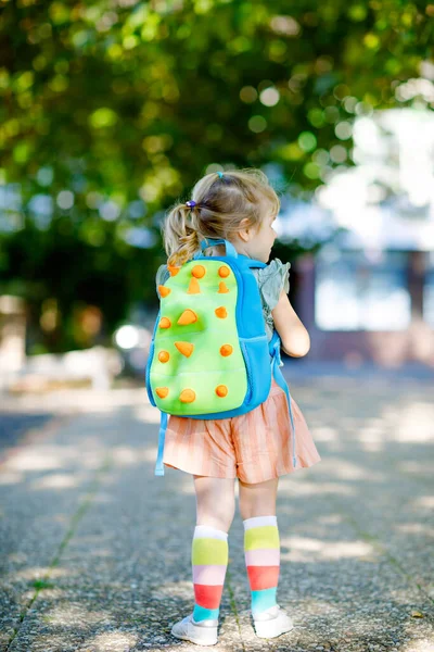 Aranyos kis aranyos kislányka az első napján, aki játszóiskolába jár. Egészséges gyönyörű baba sétál óvodába és óvodába. Boldog gyerek hátizsákkal a város utcáján, szabadban. — Stock Fotó