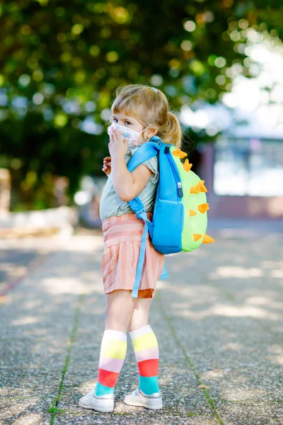 Little toddler girl on her first day on way to playschool with medical mask against corona covid virus. Healthy beautiful baby walking to nursery preschool and kindergarten. Happy child with backpack — Stock Photo, Image