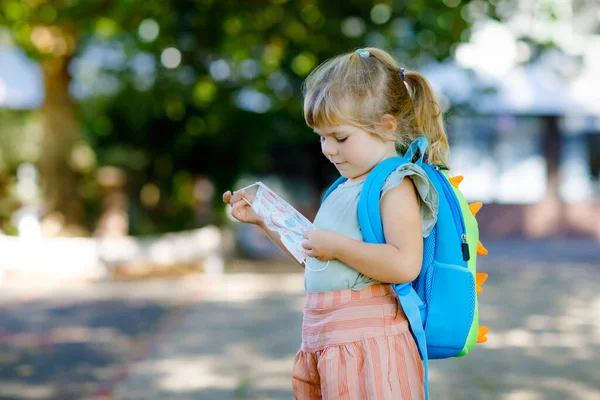 Kleines Kleinkind an ihrem ersten Schultag mit medizinischer Maske gegen Coronavirus. Gesunde schöne Baby zu Fuß in die Vorschule und den Kindergarten. Glückliches Kind mit Rucksack — Stockfoto