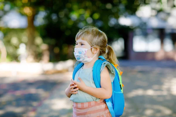 Niña pequeña en su primer día de camino a la escuela de juego con máscara médica contra el virus de la corona covid. Saludable hermoso bebé caminando a la guardería preescolar y jardín de infantes. Niño feliz con mochila — Foto de Stock