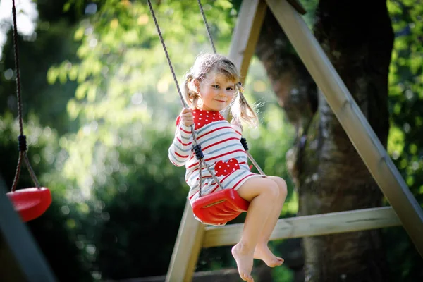 Feliz hermosa niña pequeña divirtiéndose en el columpio en el jardín doméstico. Lindo niño sano balanceándose bajo árboles florecientes en el soleado día de primavera. Bebé riendo y llorando —  Fotos de Stock