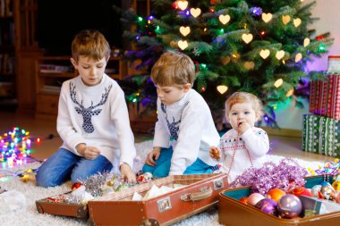 İki küçük oğlan ve sevimli kız bebek Noel ağacını eski model oyuncaklar ve toplarla süslüyorlar. Aile tatili için aile hazırlık kutlaması. Evde üç çocuk, erkek ve kız kardeş var..