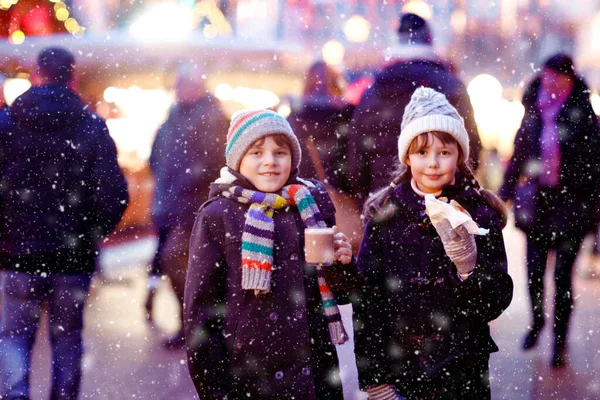 Cute little kids girl and boy having fun on traditional Christmas market during strong snowfall. Happy children eating traditional curry sausage called wurst and drinking hot chocolate. Twins friends — Stock Photo, Image