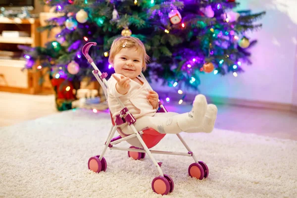 Cute adorable baby girl zabawy z wózkiem lalki. Piękne dziecko siedzące w wózku z zabawką w domu. Szczęśliwa córka. Z choinką i lampkami. — Zdjęcie stockowe