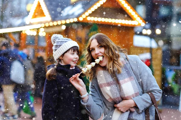 Mother and daughter eating white chocolate covered fruits and strawberry on skewer on traditional German Christmas market. Happy girl and woman on traditional family market in Germany during snowy day — Stock Photo, Image