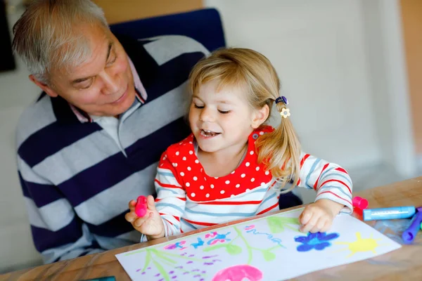 Schattig klein baby peuter meisje en knappe senior grootvader schilderij met kleurrijke vilten pennen en potloden thuis. Kleinkind en man hebben plezier samen, creatieve familie. — Stockfoto