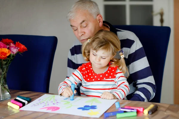 Schattig klein baby peuter meisje en knappe senior grootvader schilderij met kleurrijke vilten pennen en potloden thuis. Kleinkind en man hebben plezier samen, creatieve familie. — Stockfoto