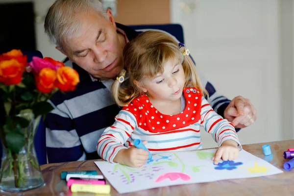 Schattig klein baby peuter meisje en knappe senior grootvader schilderij met kleurrijke vilten pennen en potloden thuis. Kleinkind en man hebben plezier samen, creatieve familie. — Stockfoto