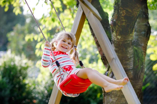 Feliz hermosa niña pequeña divirtiéndose en el columpio en el jardín doméstico. Lindo niño sano balanceándose bajo árboles florecientes en el soleado día de primavera. Bebé riendo y llorando —  Fotos de Stock