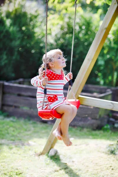 Glad vacker liten småbarn flicka som har kul på swing i hemmets trädgård. Söta friska barn svänger under blommande träd på soliga vårdagen. Baby skrattar och gråter — Stockfoto