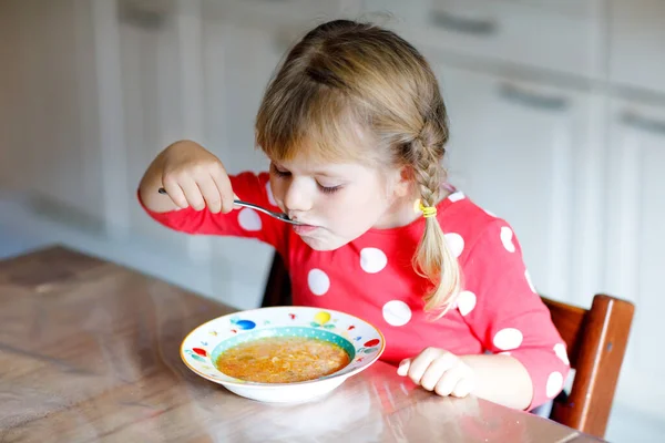 Söt liten flicka som äter färskkokt grönsakssoppa i köket. Glada barn äter hälsosam mat till lunch eller middag. Babyinlärning. Hem, förskola, lekskola eller dagis — Stockfoto