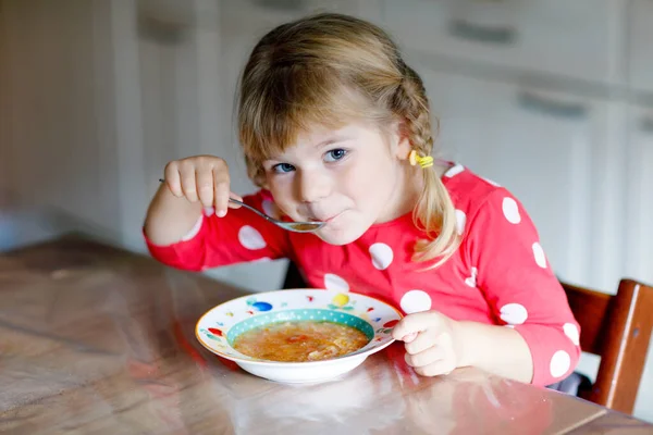 Söt liten flicka som äter färskkokt grönsakssoppa i köket. Glada barn äter hälsosam mat till lunch eller middag. Babyinlärning. Hem, förskola, lekskola eller dagis — Stockfoto