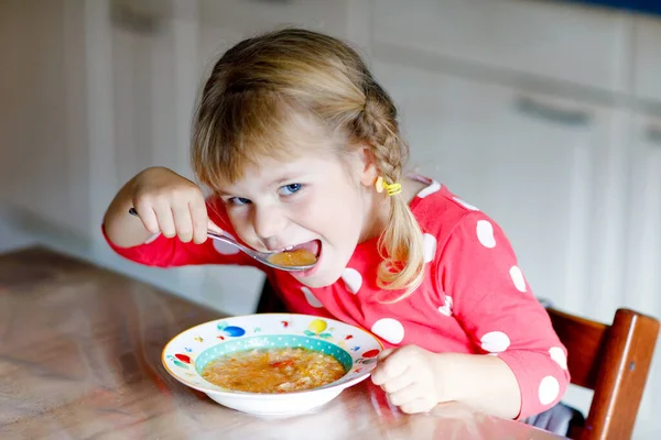 Adorável menina criança comendo sopa de legumes cozidos frescos na cozinha. Criança feliz come comida saudável ao almoço ou jantar. Aprendizagem de bebés. Casa, berçário, escola infantil ou creche — Fotografia de Stock