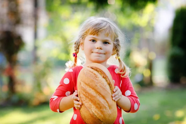 Petite fille tout-petit tenant un gros pain. Drôle enfant heureux mordre et manger du pain sain, à l'extérieur. Enfant affamé. — Photo