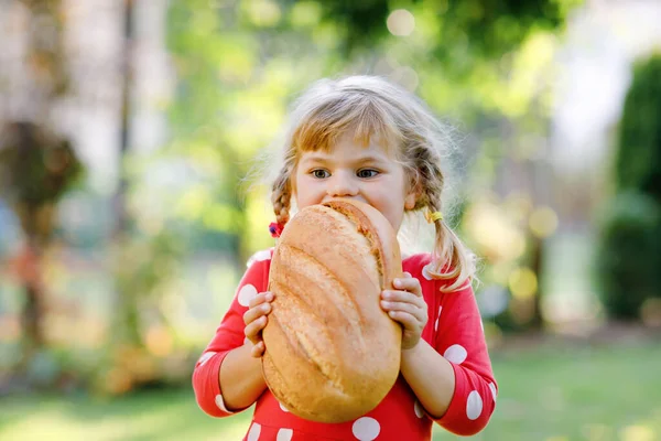 En liten flicka med en stor brödlimpa. Roligt glad barn bita och äta hälsosamt bröd, utomhus. Hungrig unge. — Stockfoto