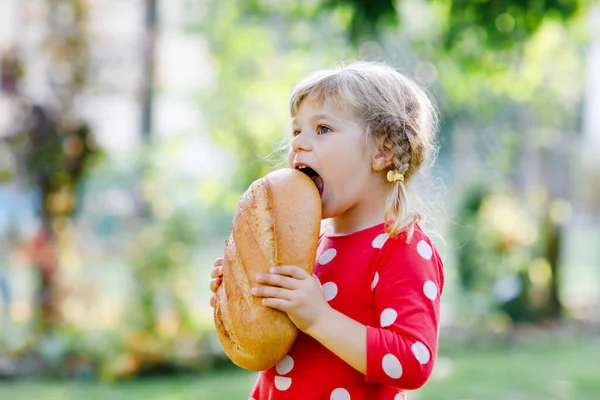 En liten flicka med en stor brödlimpa. Roligt glad barn bita och äta hälsosamt bröd, utomhus. Hungrig unge. — Stockfoto