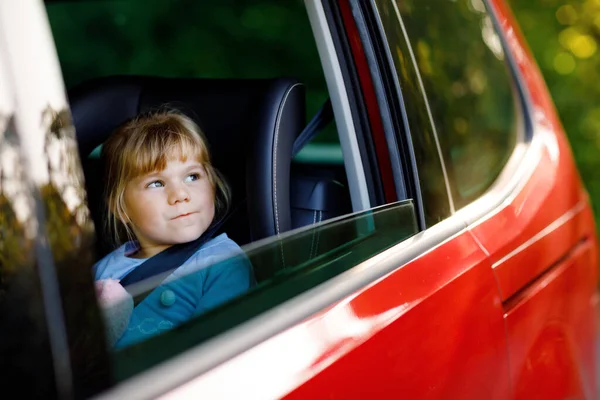 Imádnivaló kisbaba lány ül a gyerekülésen, kezében plüss puha játék, és kinézett az ablakon a természet és a forgalom. Egy kisgyerek, aki autóval utazik. Gyermekbiztonság az úton. Családi kirándulás és nyaralás — Stock Fotó