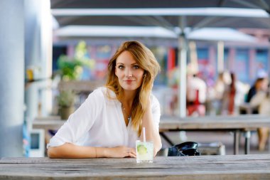 Genç bir kadın sıcak yaz gününde kafedeki terasta mojito kokteyli içiyor. Güzel bir iş kadını işten sonra restoranda sıcak bir akşam geçiriyor. Mutlu yalnız gülümseme bayan..