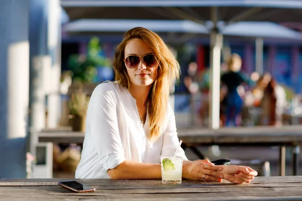 Egy fiatal nő mojito koktélt iszik a kávézó teraszán a forró nyári napon. Gyönyörű üzletasszony élvezi a meleg estét munka utáni étteremben. Boldog mosolygást, hölgyem.. — Stock Fotó