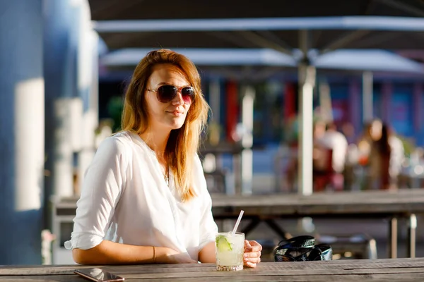 Jeune femme buvant un cocktail mojito sur la terrasse du café lors d'une chaude journée d'été. Belle femme d'affaires profitant d'une soirée chaude au restaurant afterwork. Joyeux sourire dame seule. — Photo