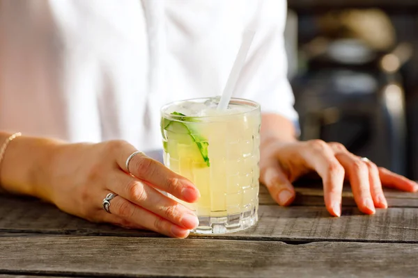 Handen van een vrouw met verse alcoholische koelcocktail in de zomer op het terras van een café. Een verfrissende mojito met ijs, munt en limoen beste idee om af te koelen in de hete zomer. — Stockfoto